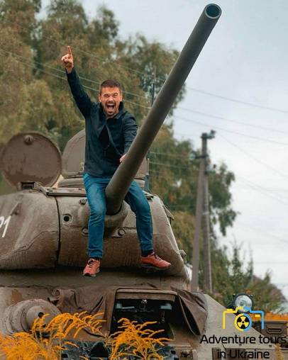 Happy man seating on the canon of a T-34 tank with the hand lifted in the air.jpg
